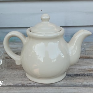 May include: Cream-colored ceramic teapot with a lid, handle, and spout. A clear glass teacup with a tea bag is to the left of the teapot. The teapot is on a weathered wooden surface.