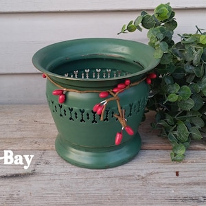May include: A green metal planter with a decorative cut-out design, adorned with a string of red berries. The planter is filled with greenery and sits on a wooden surface. The text "PineAppleBay" is visible.