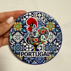 Puede incluir: Un posavasos circular con una ilustración de un gallo colorido sobre un patrón de azulejos geométricos azules y blancos. El gallo tiene una cresta roja y motivos de corazones. La palabra PORTUGAL está impresa en letras blancas mayúsculas.