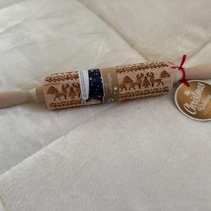 Puede incluir: Un rodillo de madera con temática navideña. El rodillo presenta diseños grabados de renos y árboles. Una etiqueta dice "Christmas Baking Products". Está atado con una cinta roja.