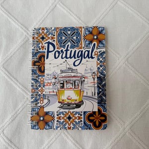 Puede incluir: Un cuaderno de espiral con una ilustración central de un tranvía amarillo y la palabra Portugal escrita encima. La portada está decorada con un borde de patrones de azulejos geométricos en tonos azul, naranja y blanco.
