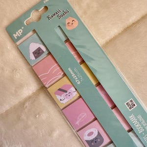 Op de afbeelding: Een pakket Kawaii Sushi plaknotities en bladwijzers. De notities bevatten kleurrijke illustraties van sushi en rijstballen. De verpakking is lichtgroen met de tekst "Kawaii Sushi" en "MP*" erop.