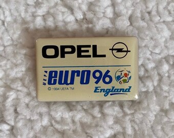 Puede incluir: Un pin de esmalte blanco y azul con el texto "UEFA EURO 96 England" y el logotipo de Opel. El pin es un objeto de colección del Campeonato de Europa de Fútbol de 1996.