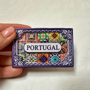 Può includere: Un magnete rettangolare viola e bianco con un motivo a piastrelle colorate e la parola "PORTUGAL" in lettere bianche.