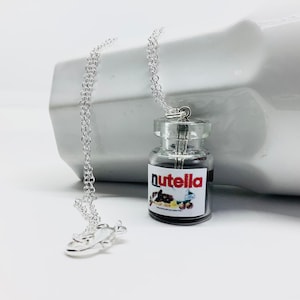 Peut inclure: Un collier en chaîne argentée avec un pendentif en forme de bocal en verre. Le bocal est rempli d'un liquide brun foncé et porte une étiquette blanche sur laquelle est inscrit "Nutella".
