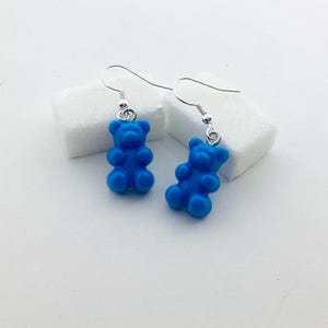 Può includere: Un paio di orecchini a forma di orsetti gommosi blu. Ogni orecchino presenta un piccolo ciondolo a forma di orsetto gommoso blu sospeso a un gancio argentato. Gli orecchini sono esposti su una superficie bianca.
