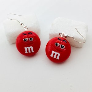 Peut inclure: Une paire de boucles d'oreilles fantaisie rouges en forme de bonbons M&M's. Chaque boucle d'oreille représente un bonbon rouge avec un "m" blanc et un visage de dessin animé. Les boucles d'oreilles ont des fermoirs en forme de crochet argentés.
