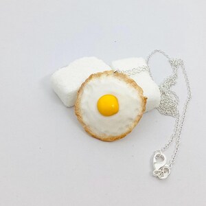 Peut inclure: Un collier argenté avec un pendentif en forme d'œuf au plat. Le pendentif a un jaune d'œuf jaune et un blanc d'œuf blanc, avec un bord marron. Le collier est à côté de deux objets blancs texturés.