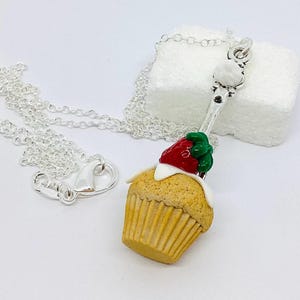 Puede incluir: Collar plateado con un colgante de cupcake. El colgante es un cupcake en miniatura con base amarilla, glaseado blanco y una fresa roja. La cadena del collar es plateada.