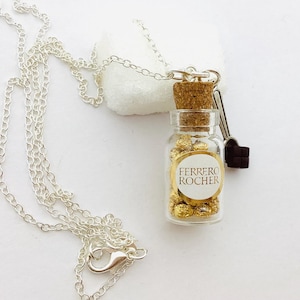 Peut inclure: Un collier en chaîne argentée avec un petit pendentif en forme de bouteille en verre rempli de chocolats enveloppés d'or. La bouteille a un bouchon en liège et une étiquette qui indique "Ferrero Rocher".