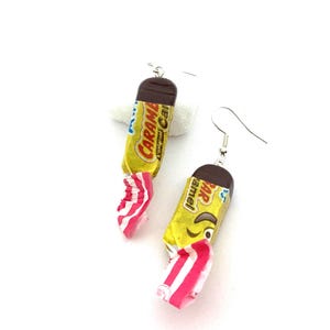 May include: A pair of earrings shaped like candy wrappers. The earrings are yellow with red and white stripes and feature the text "Caramel" and "Bar Caramel".