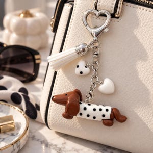 Peut inclure: Un porte-clés en métal argenté avec un fermoir en forme de cœur, un pompon blanc, deux petits cœurs blancs à pois noirs et une figurine de teckel marron portant un pull blanc à pois noirs, fixé sur un sac à main blanc.