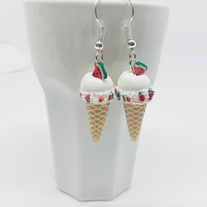 Puede incluir: Un par de pendientes de plata con un cono de helado blanco con rodajas de sandía en la parte superior. El cono de helado está hecho de arcilla polimérica y tiene un aspecto realista.