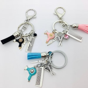 Puede incluir: Tres llaveros con herrajes plateados y dijes relacionados con el estilismo. Cada llavero presenta un borla de diferente color: negro, rosa y azul. Los dijes incluyen secadores de pelo en miniatura, tijeras, peines y un acento de cristal.