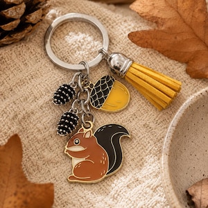Peut inclure: Un porte-clés en métal argenté composé d'une breloque écureuil marron et noir, d'une breloque gland jaune, de deux petites breloques pommes de pin noires et d'un pompon jaune aspect daim. Les breloques sont fixées à un anneau brisé argenté.