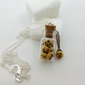 Peut inclure: Un collier argenté avec un petit pendentif en forme de bouteille en verre rempli de mini cookies aux pépites de chocolat. Une petite cuillère avec un cookie est à côté de la bouteille. Fond blanc.