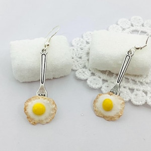 Pode incluir: Um par de brincos de novidade com um design de ovo frito. Cada brinco tem um topo em forma de garfo prateado com uma gema amarela e uma clara de ovo branca e castanha. Os brincos são apresentados numa superfície branca.