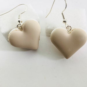 Puede incluir: Un par de pendientes en forma de corazón de color beige claro. Los pendientes están hechos de arcilla polimérica y tienen ganchos plateados.