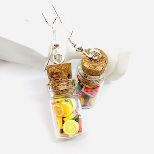 Puede incluir: Un par de pendientes con frascos de vidrio en miniatura llenos de rodajas de fruta de colores. Los frascos tienen tapas de corcho y están unidos a ganchos de pendientes de plata.