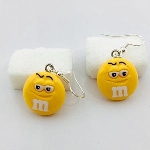 Peut inclure: Une paire de boucles d'oreilles fantaisie jaunes. Chaque boucle d'oreille présente un charm rond jaune avec un "m" blanc et un visage de dessin animé avec des sourcils et des yeux noirs. Les boucles d'oreilles ont des fermoirs en forme de crochet argentés.
