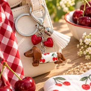 May include: A silver-toned metal keychain featuring a brown dachshund figurine wearing a white sweater with a red cherry pattern. The keychain includes two small red heart charms and a white faux leather tassel, all attached to a circular ring.