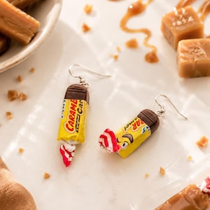 May include: A pair of dangle earrings shaped like miniature yellow and brown candy wrappers with the text Caramel printed on the front. Each earring features a silver-toned metal hook and a small, twisted red and white end detail.