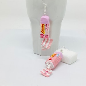 May include: A pair of novelty earrings shaped like pink and white wrapped sweets. One earring is complete, the other partially unwrapped. The wrappers feature text and a strawberry design. The earrings hang against a white backdrop.