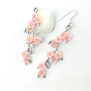 Könnte beinhalten: Ein Paar silberne Ohrringe mit rosa Blütenakzenten. Die Blüten haben gelbe Zentren und sind auf einem silbernen Zweig angeordnet.
