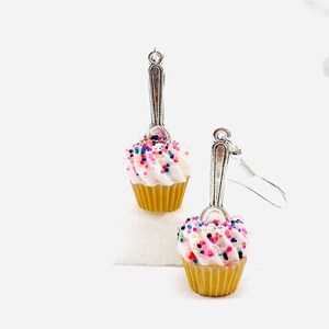 Puede incluir: Un par de pendientes de cupcake plateados con chispas rosas, blancas y azules. Los cupcakes están hechos de arcilla polimérica y tienen un aspecto realista.