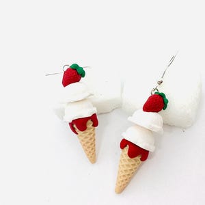 Peut inclure: Une paire de boucles d'oreilles pendantes en forme de cornet de glace blanc avec une fraise rouge et de la sauce rouge, sur fond blanc.