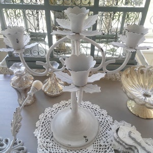 Peut inclure: Un chandelier en métal blanc avec cinq bougeoirs en forme de fleur. Le chandelier est posé sur un napperon en dentelle blanche. D'autres objets décoratifs incluent des bouteilles en verre et un bol décoratif.