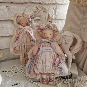 Peut inclure: Deux poupées en tissu faites à la main, ornées de fleurs et vêtues de robes vintage. L'une est une décoration suspendue, l'autre est une poupée debout. Une figurine de cheval blanc et un collier de perles sont également présents.