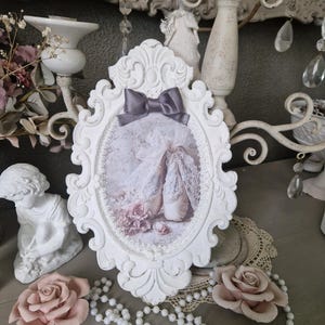 Peut inclure: Un cadre photo blanc orné d'un nœud gris, présentant une image sur le thème du ballet avec des chaussons et des roses. Des perles et des roses roses complètent la scène, créant une ambiance vintage.