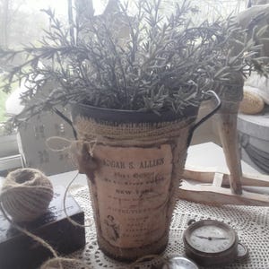 Peut inclure: Un seau décoratif en métal enveloppé de toile de jute, contenant une plante au feuillage gris-vert. Le seau a une étiquette vintage et des poignées. Une pelote de ficelle, une petite boîte et une horloge vintage sont également visibles.