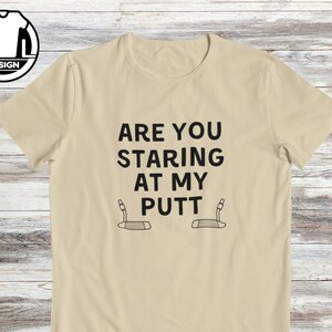 May include: A beige t-shirt with the text "Are you staring at my putt" and two golf club putters.