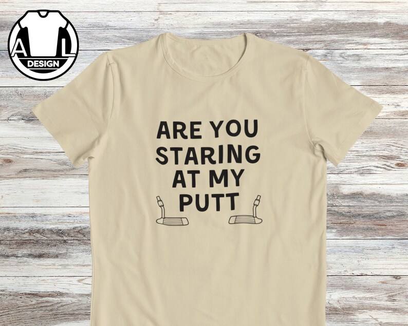 May include: A beige t-shirt with the text "Are you staring at my putt" and two golf club putters.