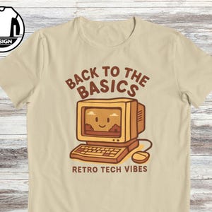 Könnte beinhalten: Beiges T-Shirt mit Retro-Computer-Grafik. Das Design zeigt einen lächelnden Computerbildschirm, eine Tastatur und eine Maus mit dem Text "BACK TO THE BASICS" und "RETRO TECH VIBES" in Braun. Das Shirt hat einen Rundhalsausschnitt und kurze Ärmel.
