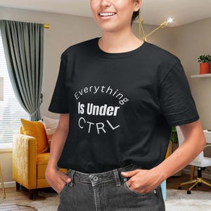 Puede incluir: Camiseta negra de cuello redondo con el texto "Everything Is Under CTRL" en un diseño circular blanco. La camiseta está hecha de un material suave y es adecuada para ropa casual. La modelo lleva vaqueros grises oscuros.