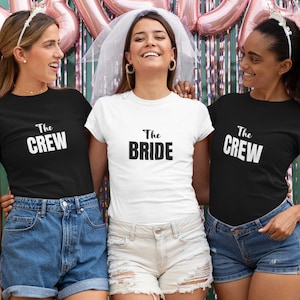 Pode incluir: Tr&ecirc;s pessoas vestindo camisetas pretas e brancas com o texto "The Crew" e "The Bride". A pessoa do meio est&aacute; vestindo uma camiseta branca e um v&eacute;u. As pessoas de cada lado est&atilde;o vestindo camisetas pretas. Todas est&atilde;o sorrindo e olhando para a c&acirc;mera.