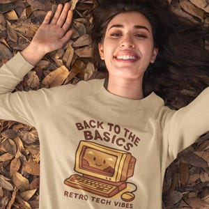 Puede incluir: Sudadera beige de manga larga con un gráfico de ordenador retro. El gráfico incluye una pantalla de ordenador sonriente, un teclado y el texto "Back to the Basics" y "Retro Tech Vibes" en marrón. La persona está tumbada sobre hojas marrones.