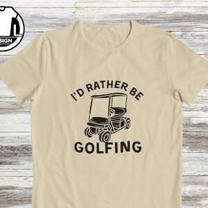 May include: A beige t-shirt with black text that reads "I'd rather be golfing" and a black silhouette of a golf cart.