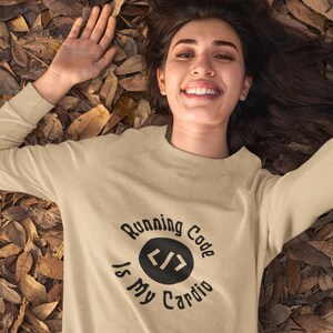 Pode incluir: Uma camisa de moletom bege com o texto "Running Code Is My Cardio" em letras pretas e um círculo preto com colchetes angulares dentro.