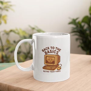 Könnte beinhalten: Weiße Keramiktasse mit einer Cartoon-Illustration eines Retro-Computers und dem Text "Back to the Basics" und "Retro Tech Vibes" in Braun. Die Tasse hat einen großen Henkel und steht auf einer Holzoberfläche.