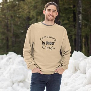Op de afbeelding: Een beige sweatshirt met de tekst "Everything Is Under CTRL" in een cirkelvormig ontwerp. De longsleeve crew neck shirt wordt gedragen door een persoon die voor een besneeuwde achtergrond staat.