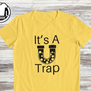 May include: Yellow t-shirt with black text that reads "It's A U Trap" with a black horseshoe-shaped graphic with white dots inside.