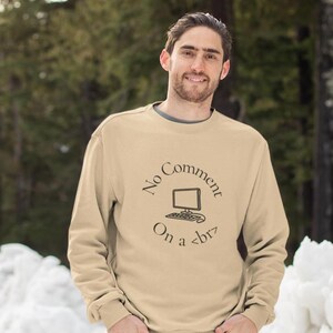 Op de afbeelding: Beige sweatshirt met de tekst "No Comment On a <br>" en een computerafbeelding. De sweatshirt heeft een ronde hals en lange mouwen. De persoon op de foto lacht, tegen een besneeuwde achtergrond.