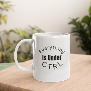 Peut inclure: Tasse à café en céramique blanche avec une anse incurvée. Le texte "Everything Is Under CTRL" en noir est imprimé sur le devant. La tasse est posée sur une surface en bois, avec des plantes en arrière-plan.