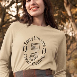 Peut inclure: Un t-shirt beige à manches longues avec un motif graphique noir qui dit "Désolé, je suis en retard, j'étais en train de coder" avec un écran d'ordinateur, du code et des crochets.