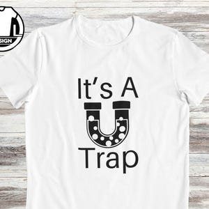 May include: White t-shirt with black text that reads "It's A U Trap" with a black horseshoe-shaped magnet with white dots.