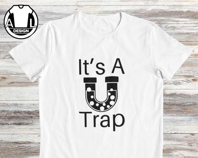 May include: White t-shirt with black text that reads "It's A U Trap" with a black horseshoe-shaped magnet with white dots.